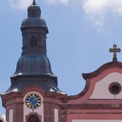 Turm einer Kirche mit einer blauen Uhr und einer Kreuzspitze mit rosa und roten Fassadenelemente.