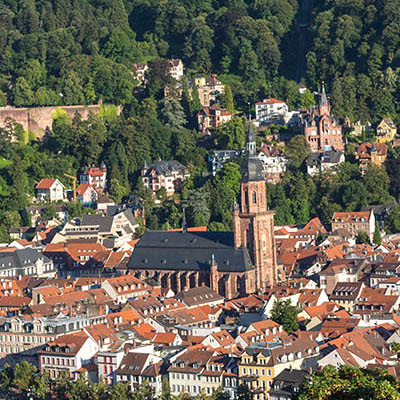Luftaufnahme von Heidelberg mit roten Dächern, einer Kirche mit hohem Turm und umgebenden grünen Hügeln.