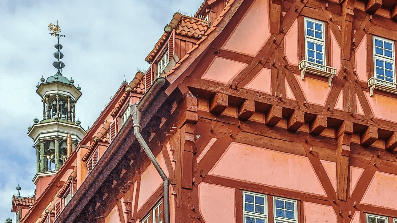 Detailansicht eines historischen Fachwerkhauses mit rosa Fassade und Holzverzierungen, im Hintergrund ein Turm mit Wetterfahne.