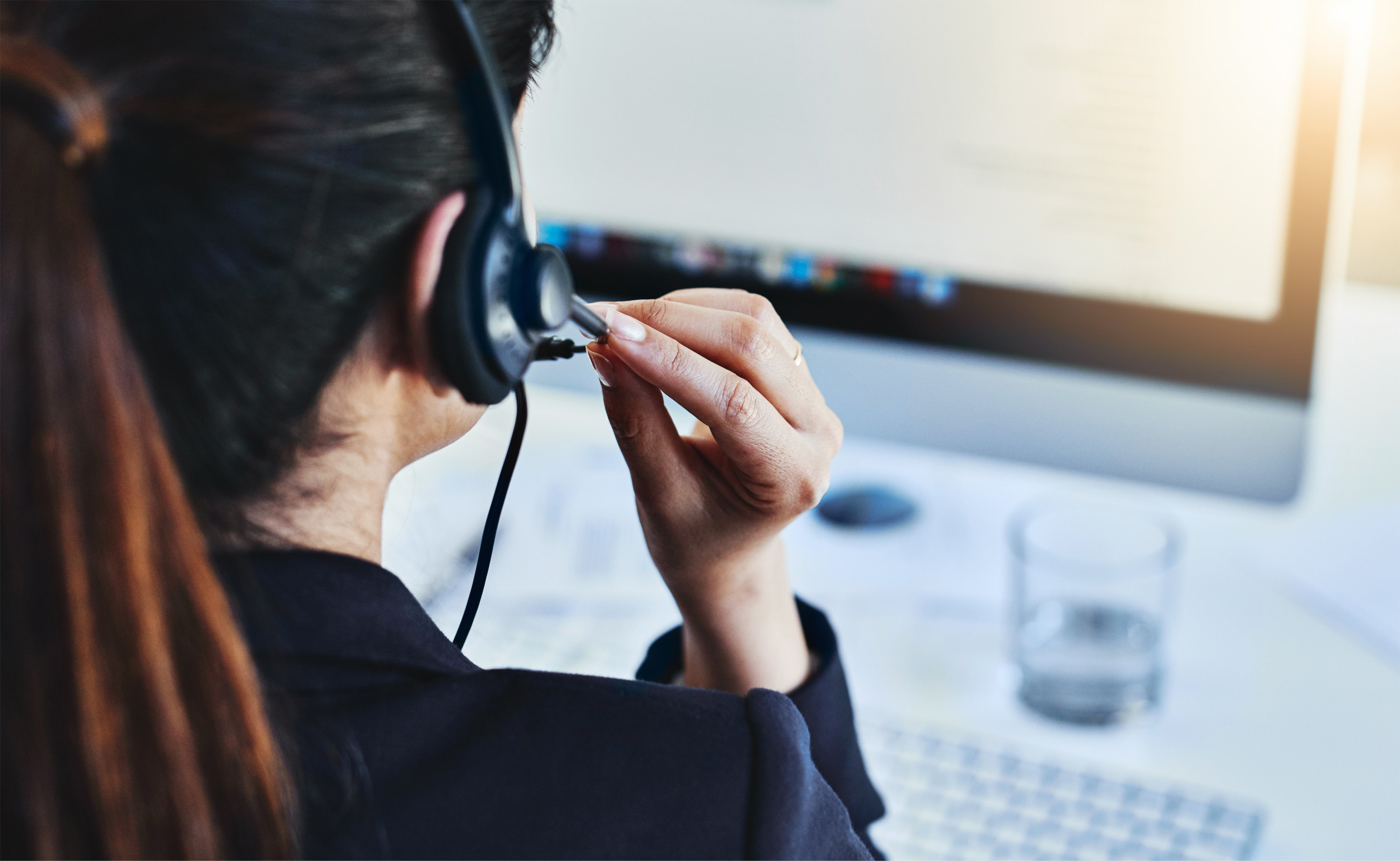 Eine Person mit Headset auf dem Kopf, sitzend vor einem Arbeitsplatz, Monitor, Tastatur und ein Wasser vor sich stehend.