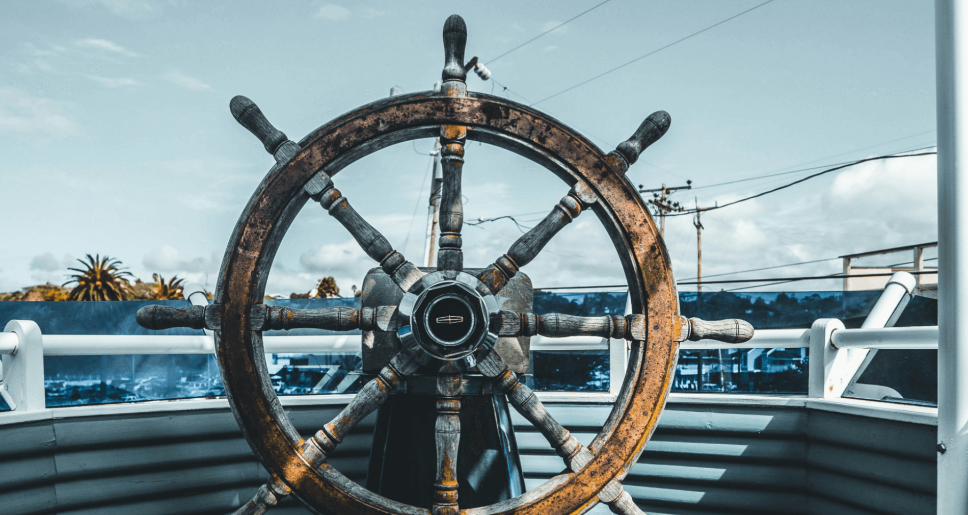 Schiffsrad aus Holz mit Metallgriffen, im Hintergrund eine Küstenlandschaft und Stromleitungen.