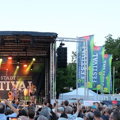 Bühne auf dem 3-Länder-Stadt-Festival mit Musikband und einer Menschenmenge davor. Rechts daneben stehen drei Fahnen mit dem Logo des Festivals.