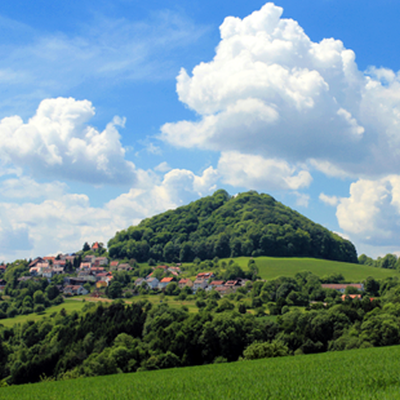 Grüne Hügel mit bewaldetem Gipfel unter einem blauen Himmel mit weißen Wolken, im Vordergrund ein kleines Dorf im Landkreis Göppingen.