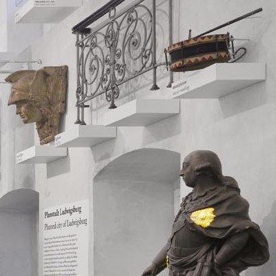 Historische Exponate in einem Museum: Eine Statue eines Mannes, ein antikes Trommelinstrument und eine maskenartige Skulptur auf Regalen.