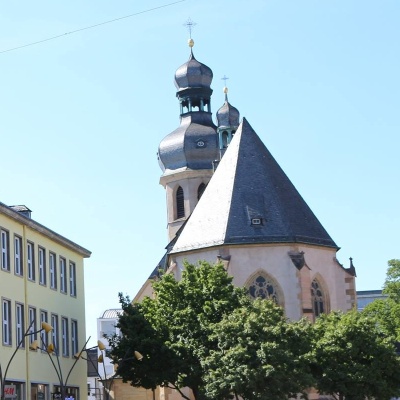 Dach und zwei Türme einer Kirche mit filigranen Kreuzen auf den Spitzen. Davor ein grüner Baum und blauer Himmel im Hintergrund.