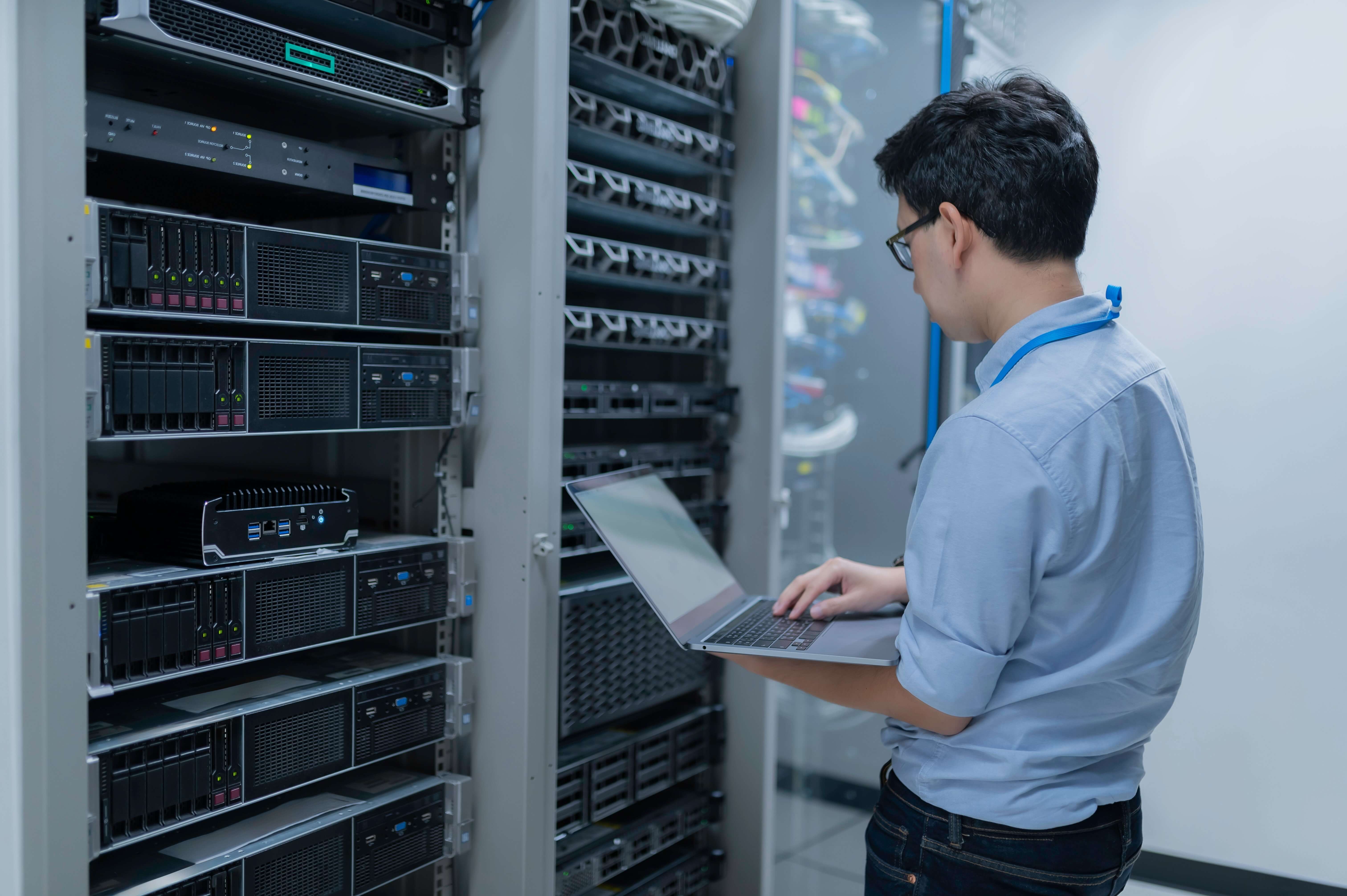 Eine Person stehend vor Servern mit Festplatten bestückt, die Person hält einein Laptop in der Hand .