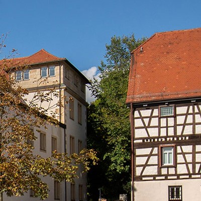 Historische Gebäude mit Fachwerkarchitektur und modernen Elementen, umgeben von Bäumen und blauem Himmel.