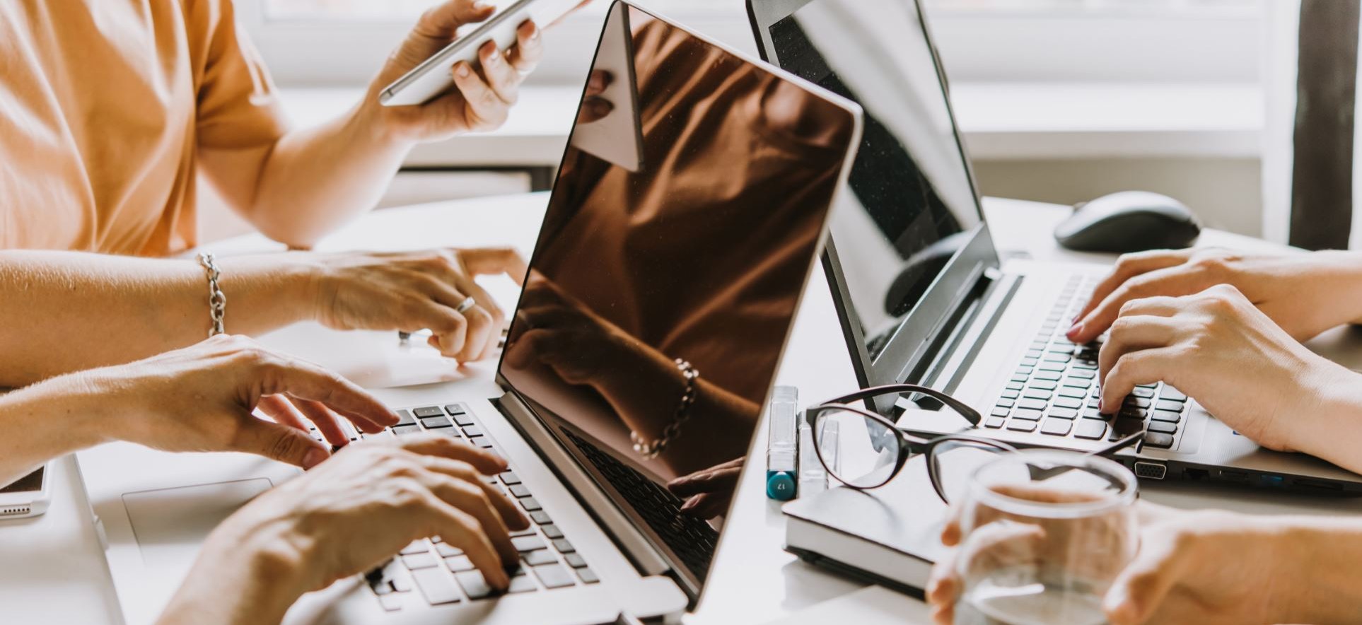 Zwei Personen arbeiten an Laptops, eine Dritte hat ein Handy in der Hand. Hände tippen auf Tastaturen, Brille liegt auf dem Tisch.