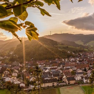 Sonnenuntergang über der Gemeinde Steinach, umgeben von Hügeln und Bäumen, mit Windrädern im Hintergrund.
