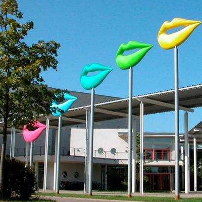 Fünf große, bunte Lippen-Skulpturen in verschiedenen Farben (pink, grün, blau, gelb) auf hohen Stangen vor einem modernen Gebäude.