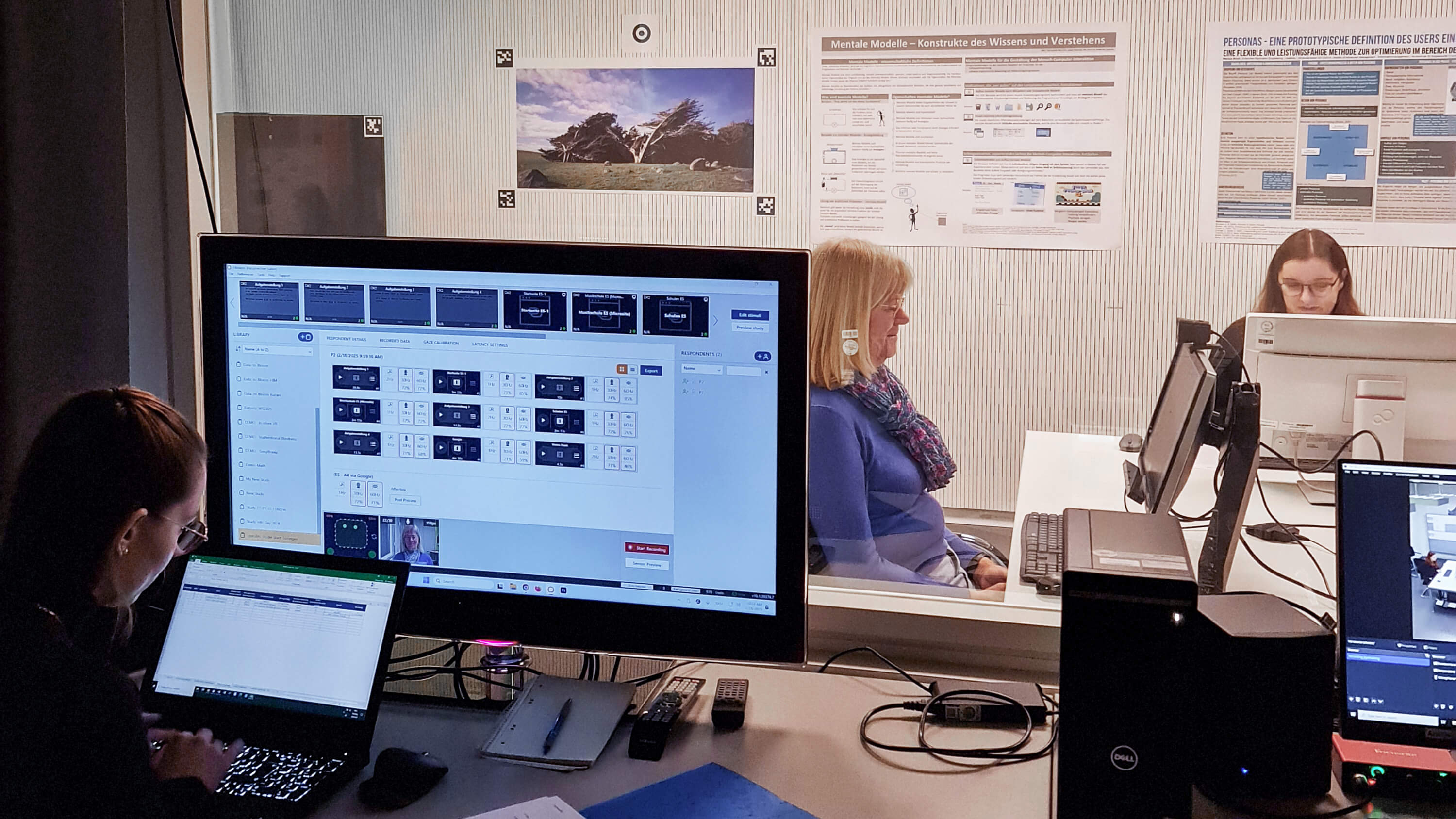 Eine große Glasscheibe, die den Raum trennt. Auf der einen Seite zwei Personen an zwei Bildschirmen, auf der anderen Seite eine Person, die sich Ergebnisse notiert.