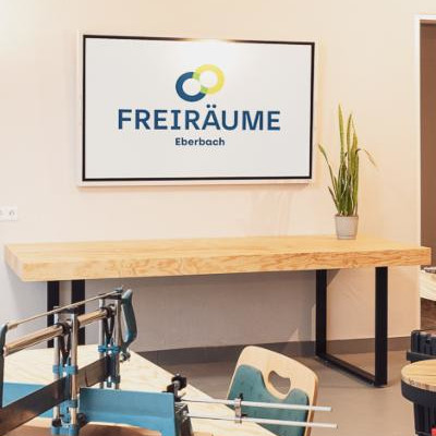 Logo von FREIRÄUME Eberbach auf einem Bildschirm an einer Wand, darunter ein Holztisch mit Pflanzen.