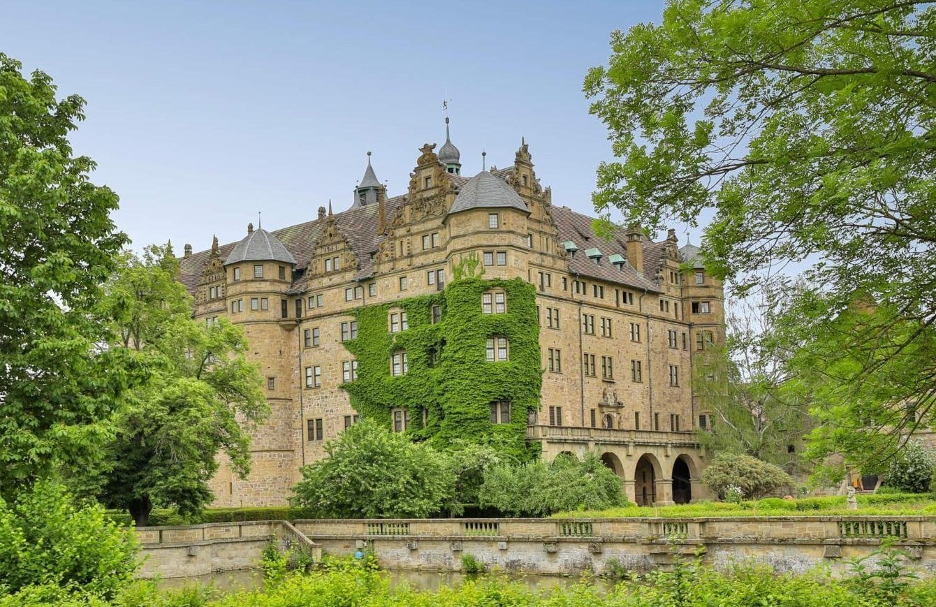 Historisches Schloss mit Türmen und grüner Bewachsung, umgeben von Bäumen und Sträuchern.