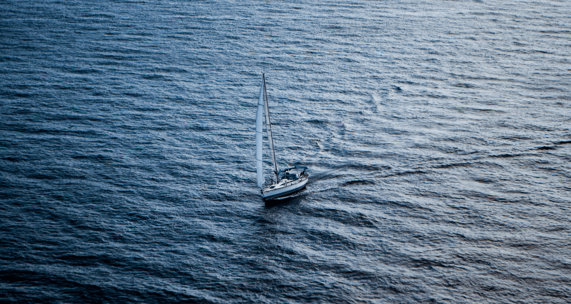 Ein Segelboot mit weißen Segeln fährt über ruhige, blaue Wasseroberfläche.