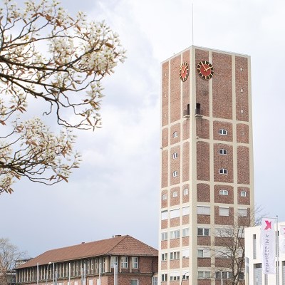 Hoher eckiger Turm aus Backsteinen mit einer roten Turmuhr.