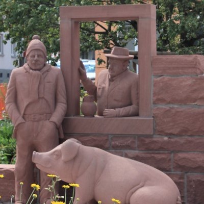 Skulpturengruppe in Malsch aus Sandstein mit zwei Männern, einer im Hut und der andere in Mütze, sowie einem Schwein.
