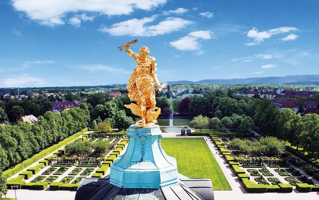 Die goldene Statue einer kriegerischen Figur mit Schwert und Schild, umgeben von einer grünen Landschaft und blauen Himmel in Rastatt.